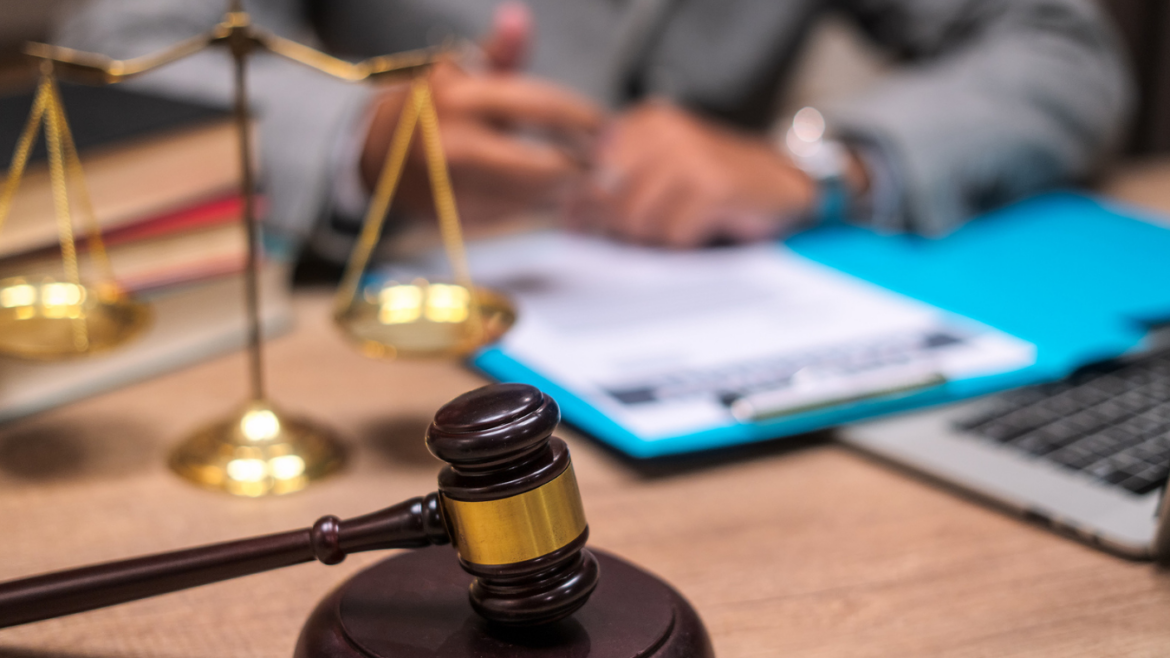 Close up shot of lawyer's desk with gavel, scales of justice, paperwork, books, and laptop. Male lawyer can be seen in background, wearing a suit and going over documents.