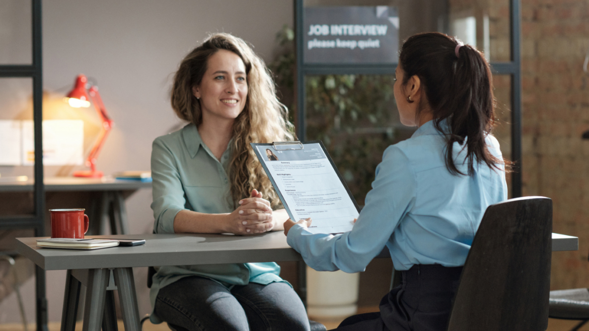 Young woman sitting across desk from female manager who is going over her application. Inside modern office.