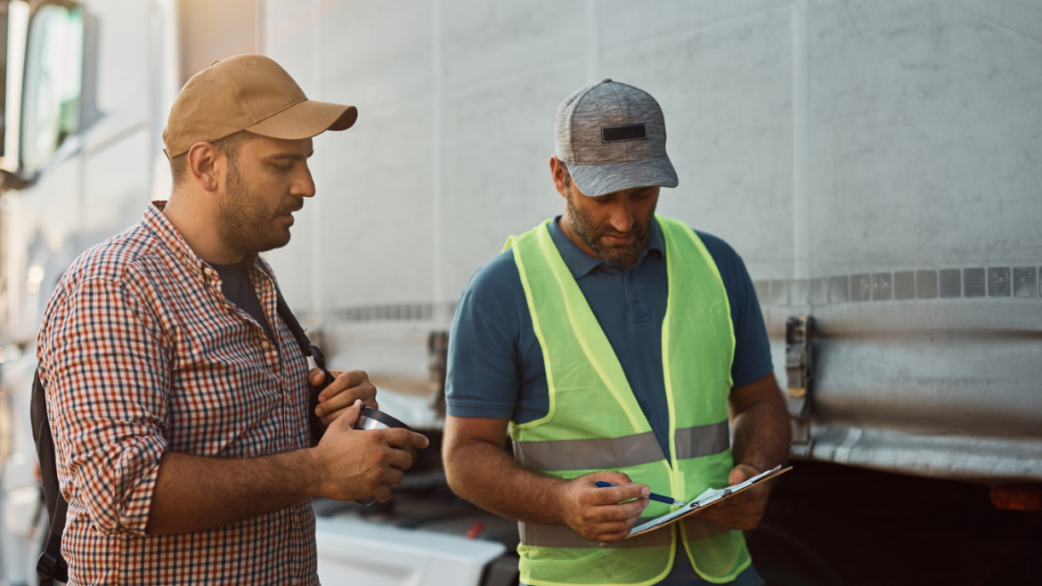 Two adult male truck drivers standing in front of truck, looking at clipboard.