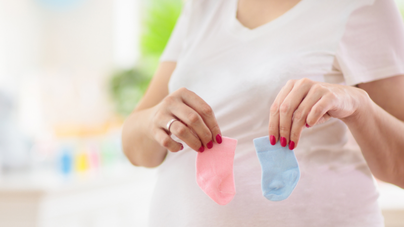 Pregnant woman in nursery holding up one pink baby sock and one blue baby sock.
