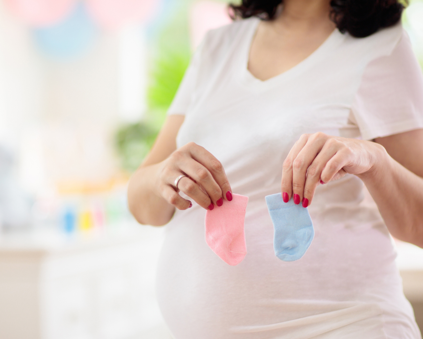 Pregnant woman in nursery holding up one pink baby sock and one blue baby sock.